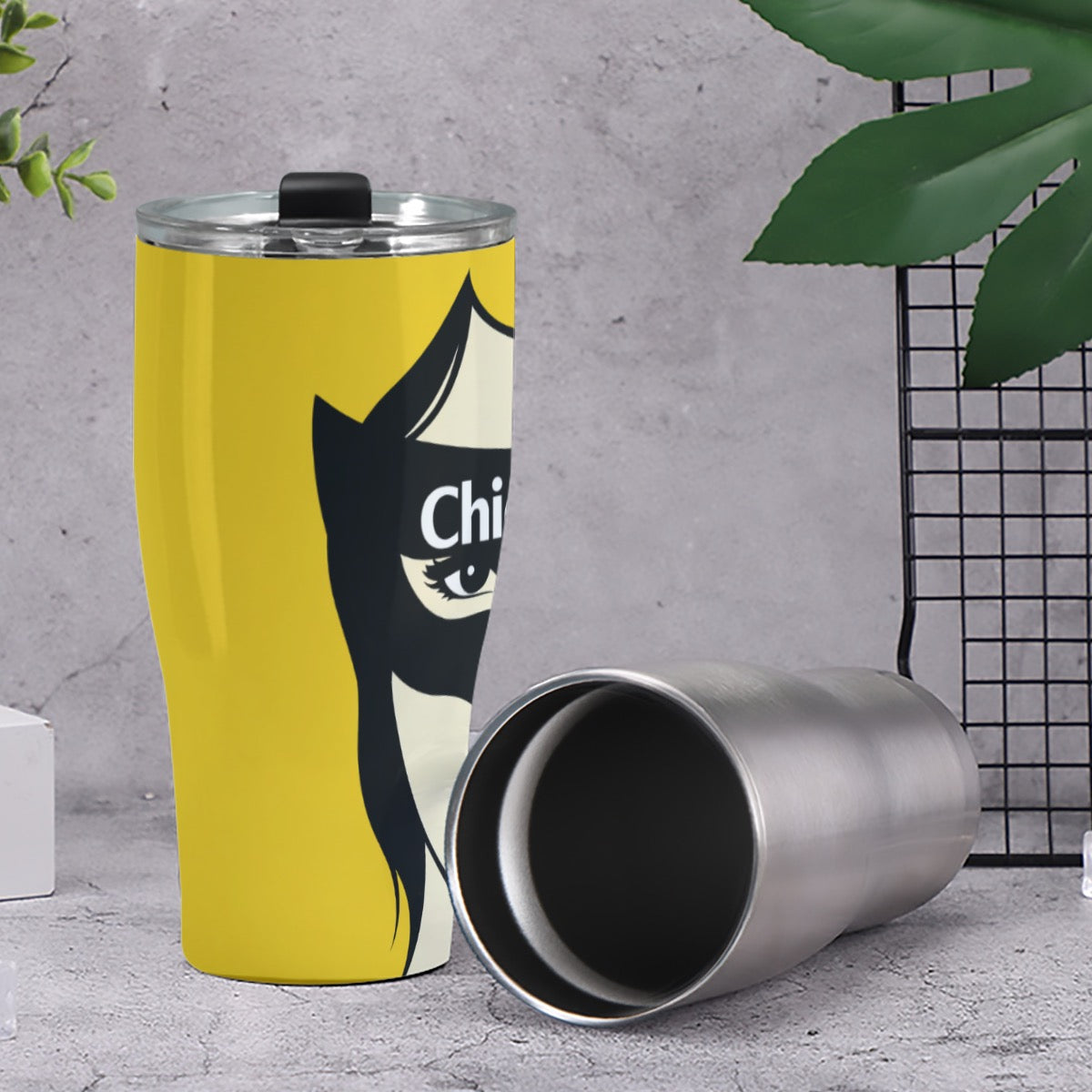 Yellow tumbler with a black and white design on a gray surface with green leaves in the background